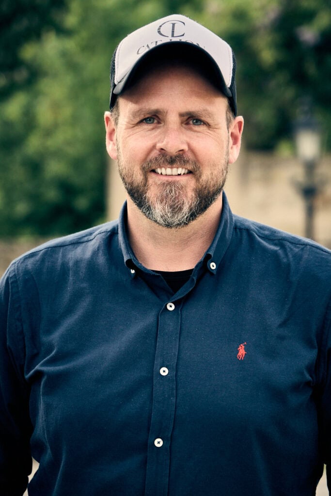 Mann im Freien, lächelnd, mit dunklem Hemd und Cap.